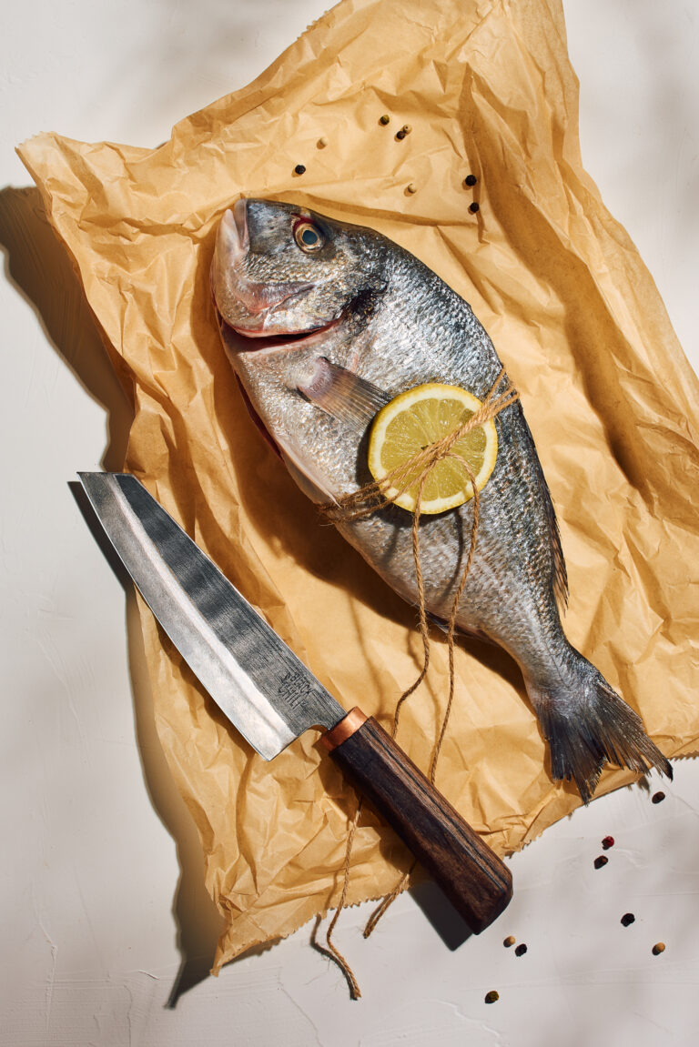 High-end product photography showcasing premium kitchen knives by Black Chili, combined with fresh ingredients to demonstrate functionality within a bold, minimalist composition, showcasing a whole raw fish wrapped in crumpled brown paper with a lemon wheel slice, positioned elegantly next to a sharp steel blade.
