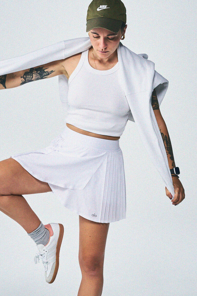 Contemporary lifestyle and product photography showcasing the iconic Samba sneaker design in a minimalist, urban fashion setting, capturing a dynamic action moment focusing on the white pleated skirt and a white sweater draped over the model's shoulders.