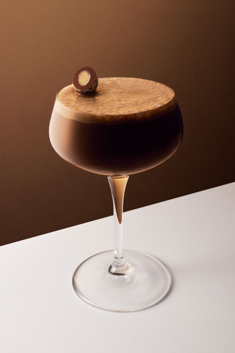 Premium liquid product photography showcasing a minimalist cocktail setup with sophisticated lighting that accentuates the glass contours and ice textures, showcasing a creamy chocolate cocktail on a rich brown background, topped with a single, perfectly centered chocolate-coated nut.