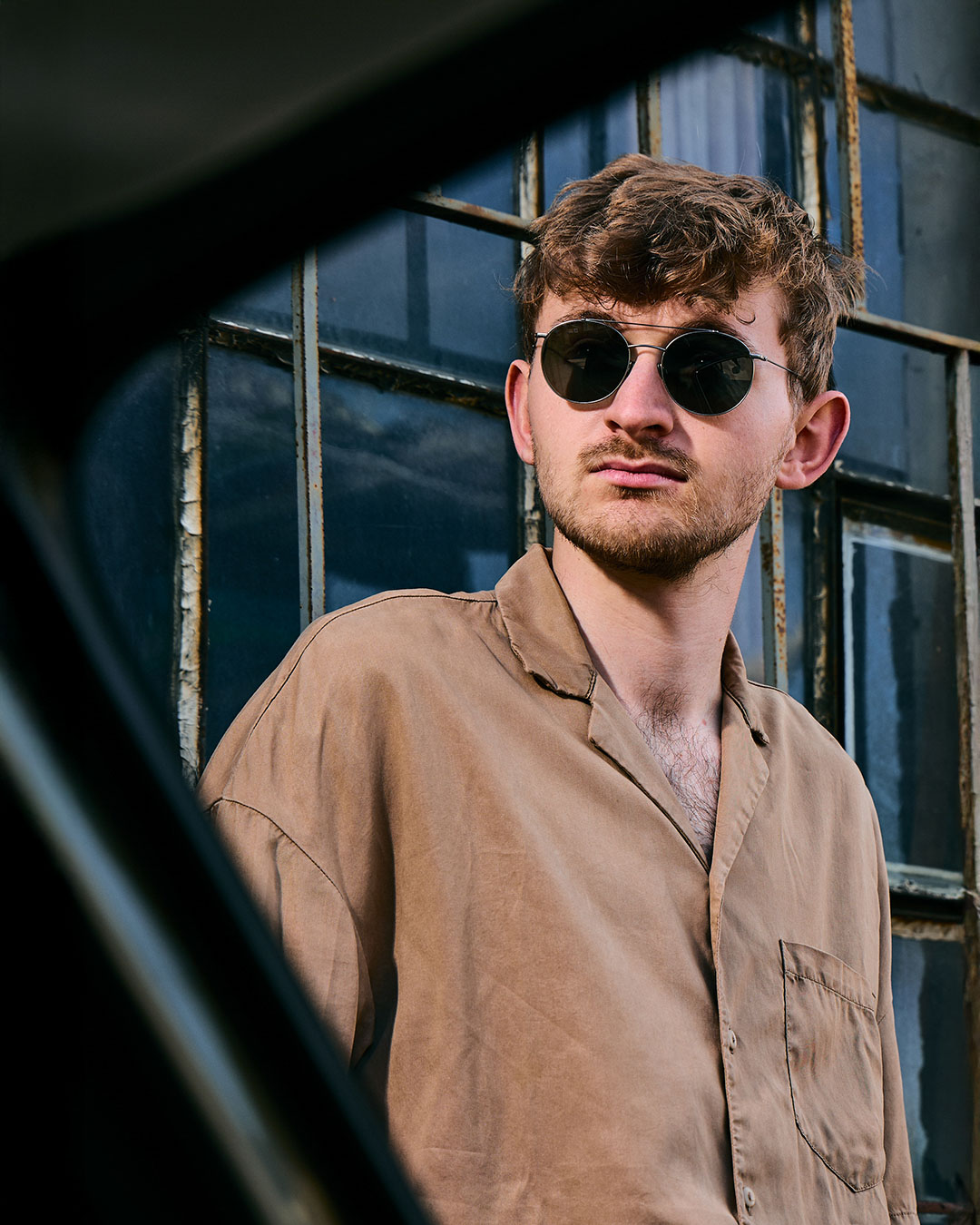 High-end outdoor editorial fashion photography by Lara Zeh featuring a model with sunglasses posing with a vintage Volkswagen Beetle in an industrial brickyard setting, featuring a sharp studio-style outdoor portrait highlighting the classic round sunglasses.