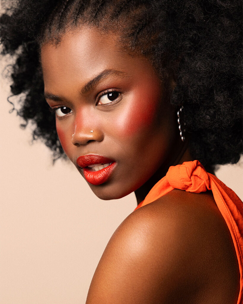 Professional beauty campaign image for Faber-Castell Cosmetics featuring clean studio portraits with diverse models and soft lighting that defines natural contours and warm earth tones, highlighting a striking beauty portrait in vibrant red styling with soft, warm edge-lighting.