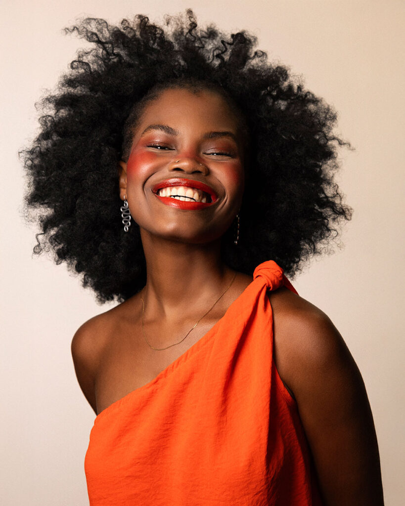 Professional beauty campaign image for Faber-Castell Cosmetics featuring clean studio portraits with diverse models and soft lighting that defines natural contours and warm earth tones, showcasing a joyful, smiling portrait that emphasizes glowing skin and radiant red lip tones.