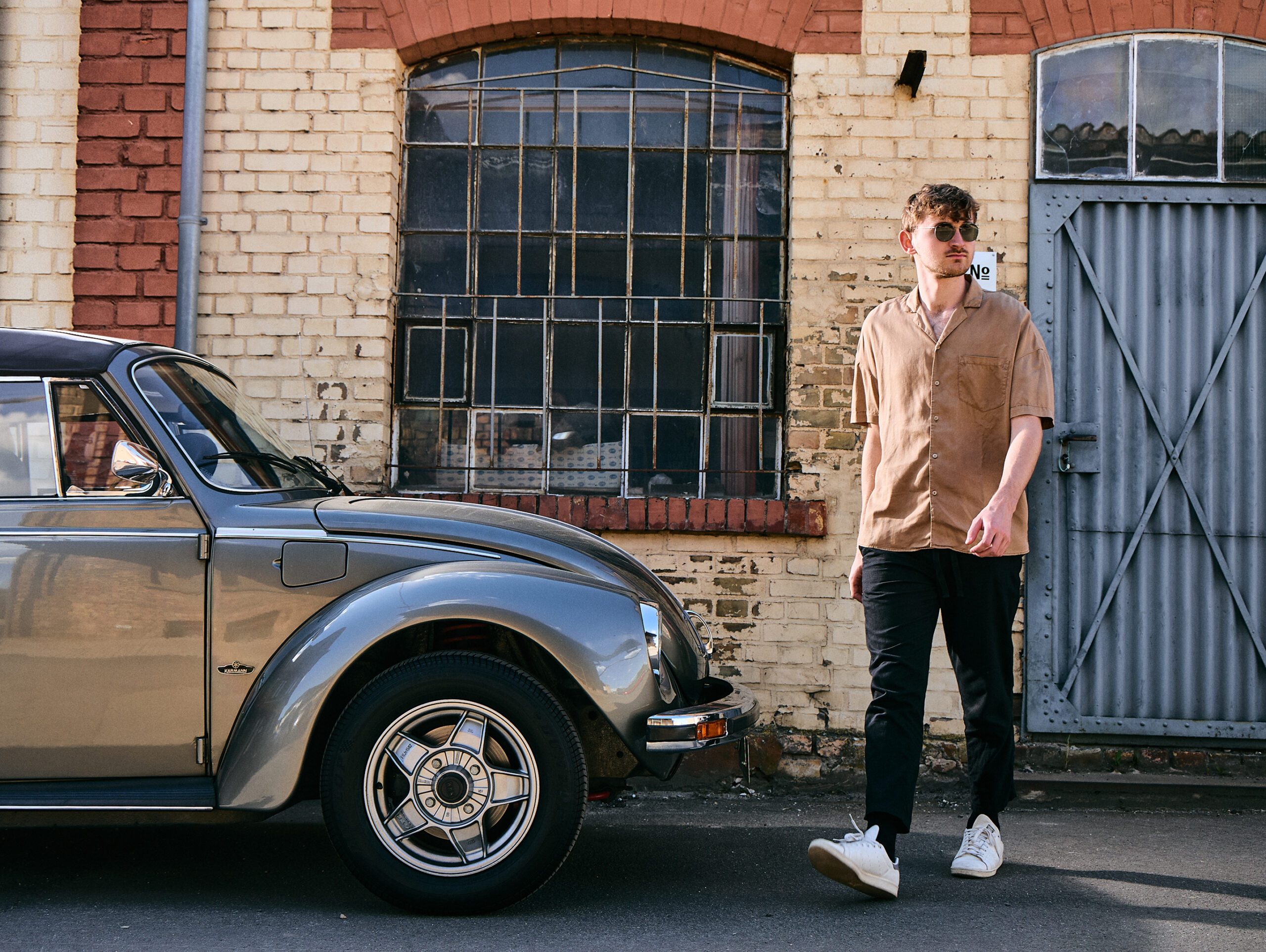 High-end outdoor editorial fashion photography by Lara Zeh featuring a model with sunglasses posing with a vintage Volkswagen Beetle in an industrial brickyard setting, showcasing a full-length lifestyle shot as the model walks past the front of the vintage car.