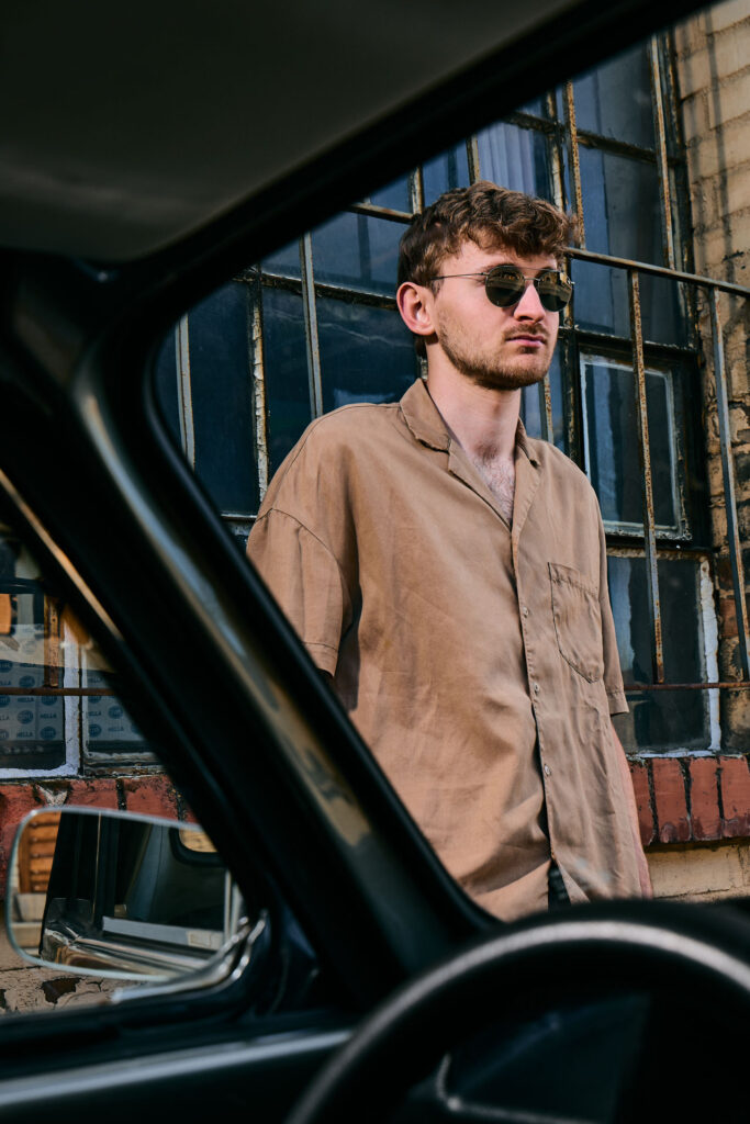 High-end outdoor editorial fashion photography by Lara Zeh featuring a model with sunglasses posing with a vintage Volkswagen Beetle in an industrial brickyard setting, capturing a medium shot framed through a car window with a grid pattern background.