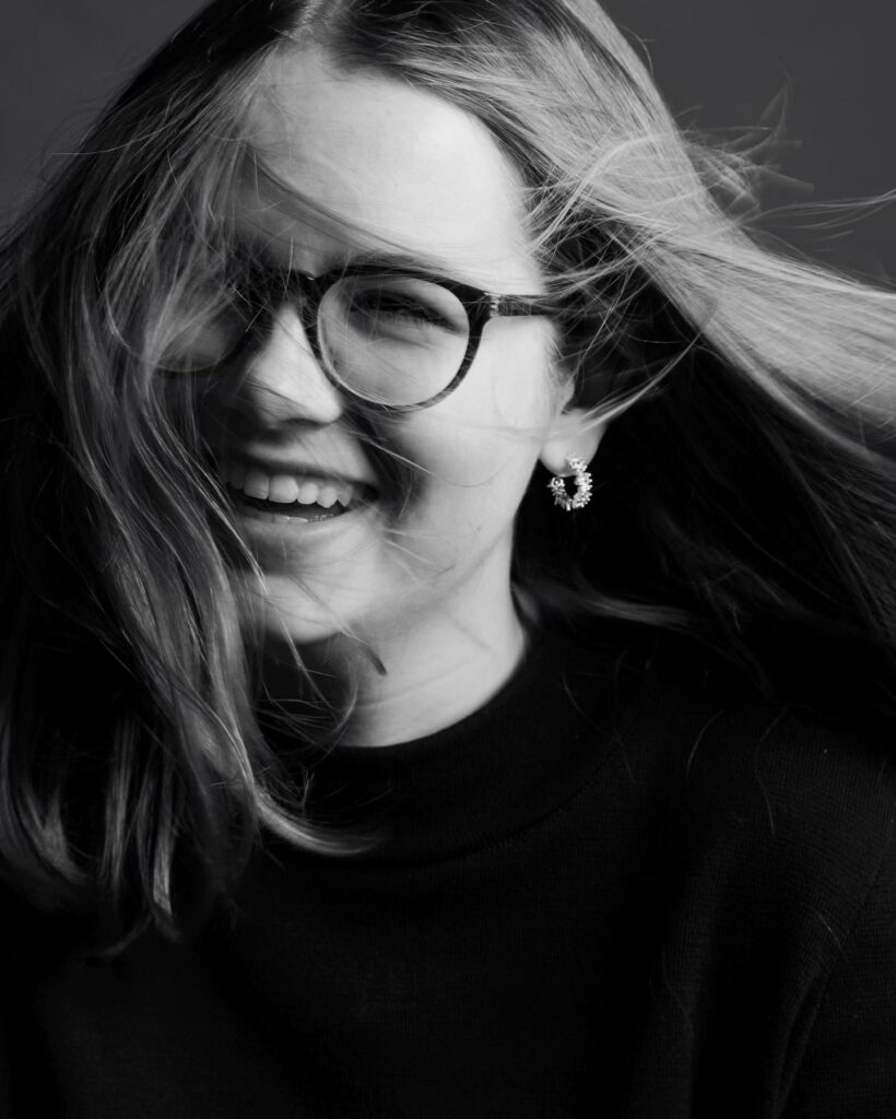 Professional black and white studio portrait of freelance photographer Lara Zeh, featuring a joyful, smiling expression with glasses and a minimalist dark sweater, representing her creative and modern brand aesthetic.