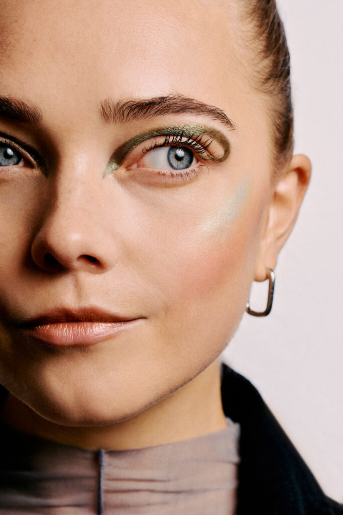 High-fashion beauty commercial shot for Faber-Castell Cosmetics featuring multiple models, capturing the sharp metallic reflection and radiance of glitter pigments on the skin under direct lighting, showcasing a tight close-up emphasizing a metallic green and silver dual-liner concept.
