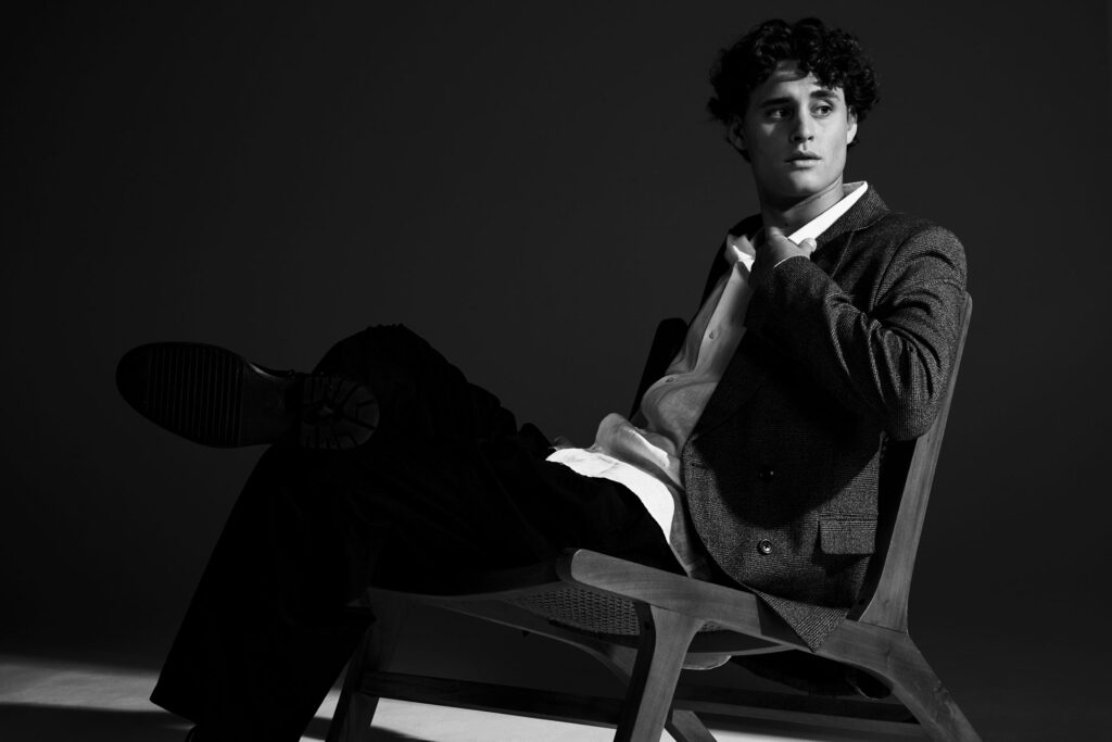 Minimalist studio fashion editorial showcasing a modern brown suit and a flat graphic wooden chair, focusing on structural geometry and high-contrast lighting, utilizing a stark black and white edit to focus on the sharp geometric shadows and dynamic body lines.