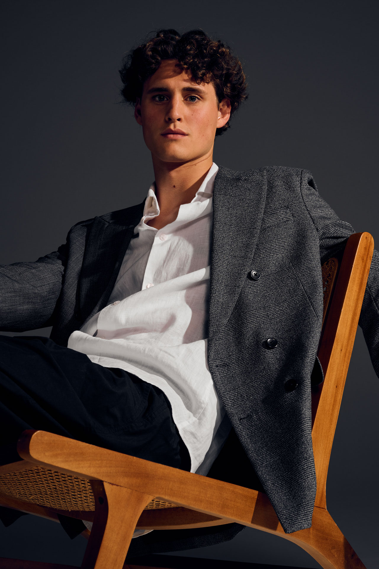 Minimalist studio fashion editorial showcasing a modern brown suit and a flat graphic wooden chair, focusing on structural geometry and high-contrast lighting, highlighting a frontal close-up that emphasizes the clean textures of the white shirt and the wooden chair frame.
